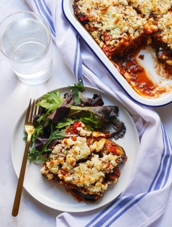 A birdseye shot of aubergine bake on a white plate with a side salad and a gold fork. The baking tray which the bake was cooked in is next to the plate with three more slices of aubergine bake. A blue and white napkin is placed in between the plate and baking tray.